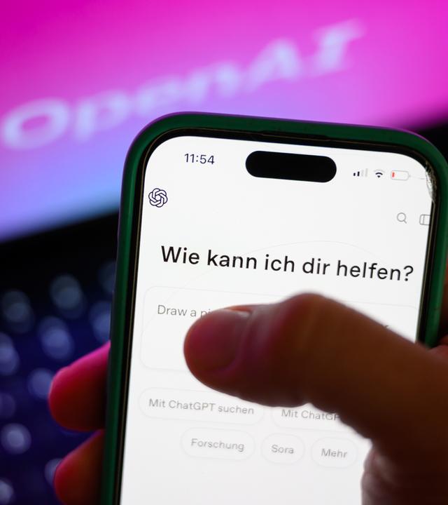 Vor einem Laptop mit Open AI-Hintergrund hält jemand ein Smartphone mit geöffneter ChatGPT-App.