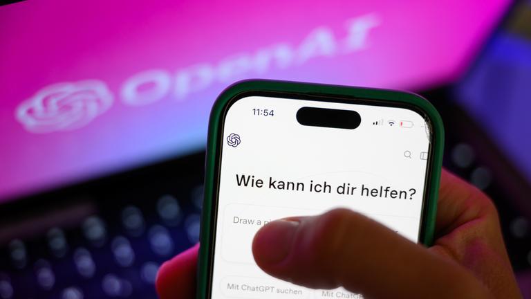 Vor einem Laptop mit Open AI-Hintergrund hält jemand ein Smartphone mit geöffneter ChatGPT-App.