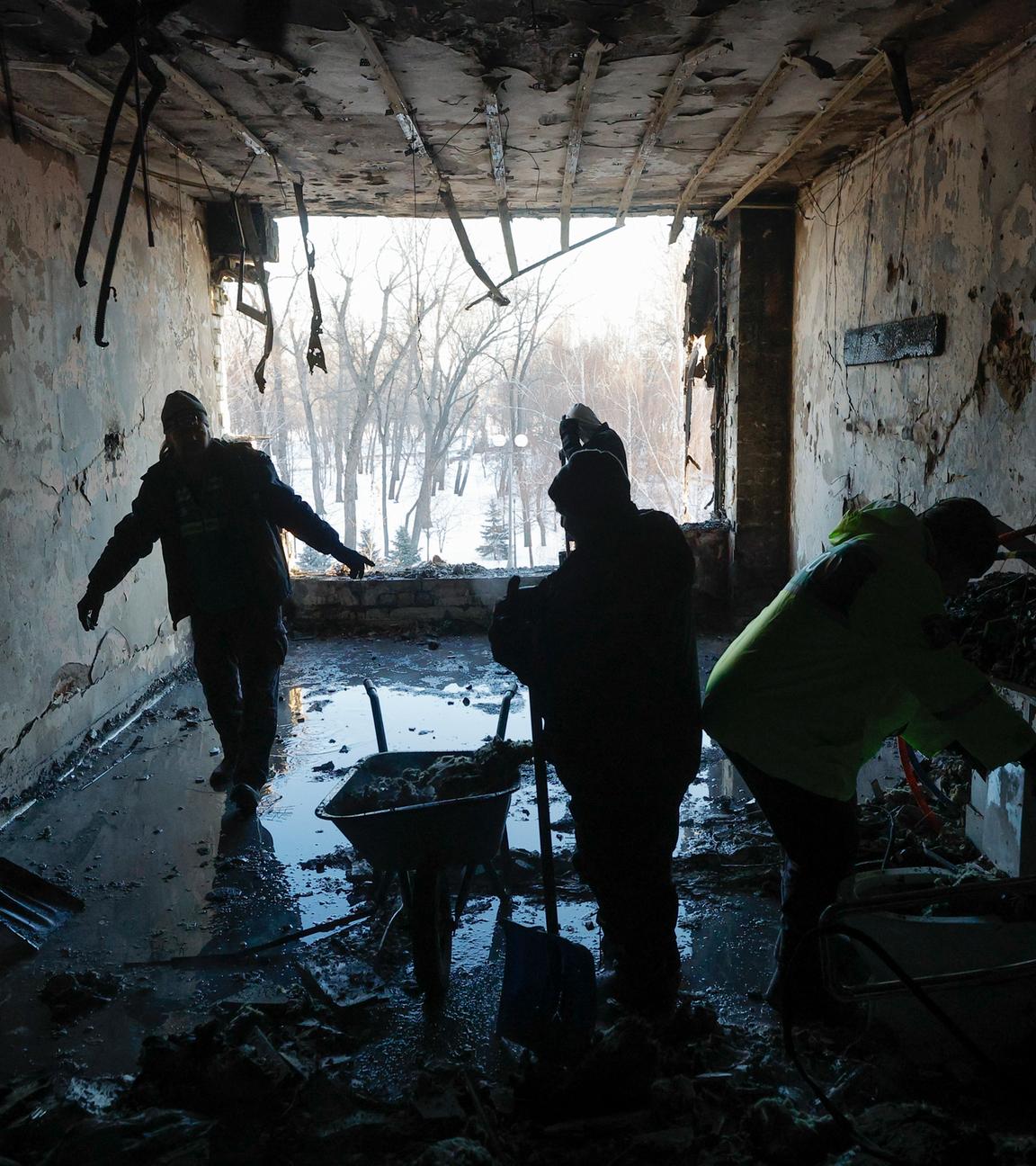 Drei ukrainische Männer räumen in einem zerstörten Gebäude Schutt weg, nasser Untergrund und eine Schubkarre sind zu sehen. Recht dunkle Szenerie.