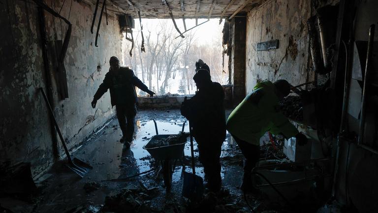 Drei ukrainische Männer räumen in einem zerstörten Gebäude Schutt weg, nasser Untergrund und eine Schubkarre sind zu sehen. Recht dunkle Szenerie.