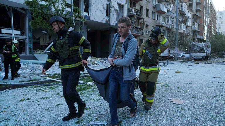 Drei Männer tragen eine verletzte Frau mit einer Trage weg. Im Hintergrund Schutt von beschädigten und zerstörten Gebäuden