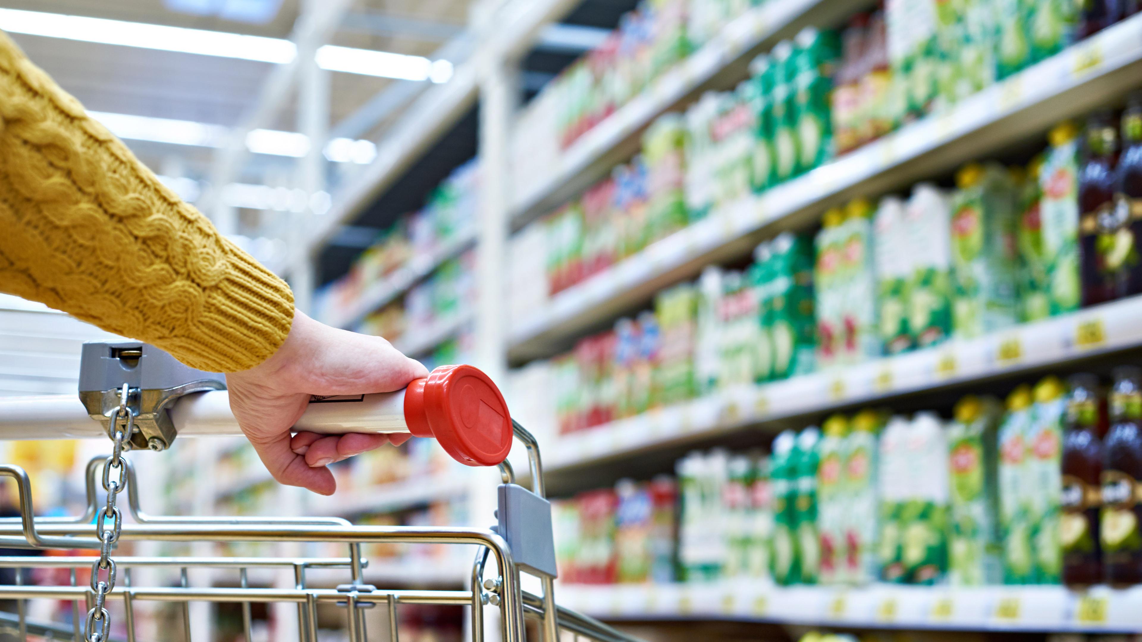 Das Bild zeigt wie eine Frau einen Einkaufswagen durch einen Supermarkt schiebt.