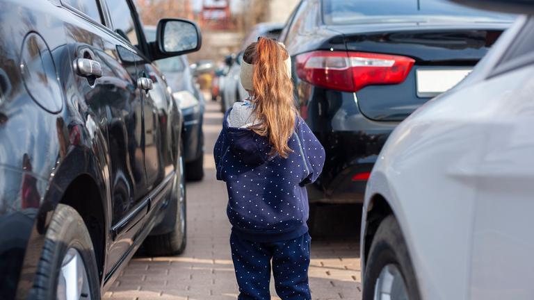 Kind steht aleine zwischen Autos auf einem Parkplatz.