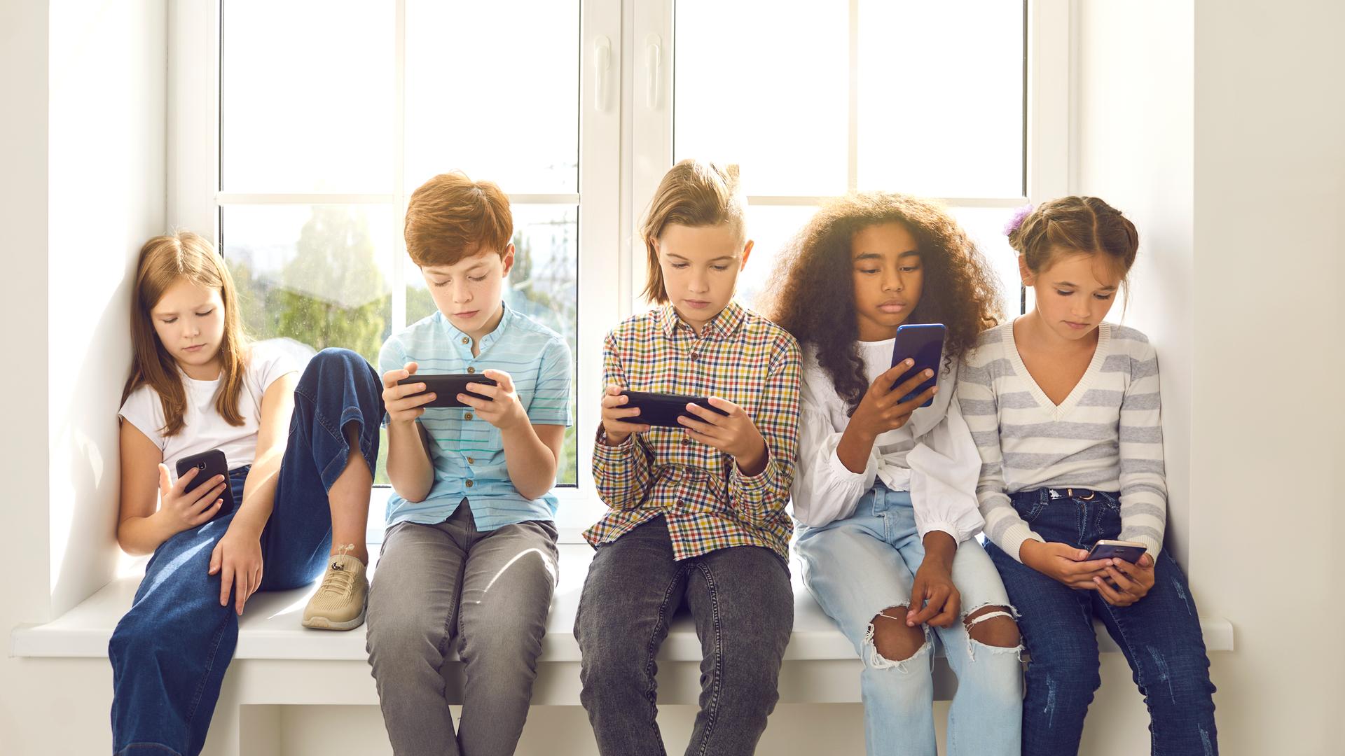 Eine Gruppe Kinder im Alter von 8-10 Jahren sitzt auf der Fensterbank und spielt Online-Handyspiele.