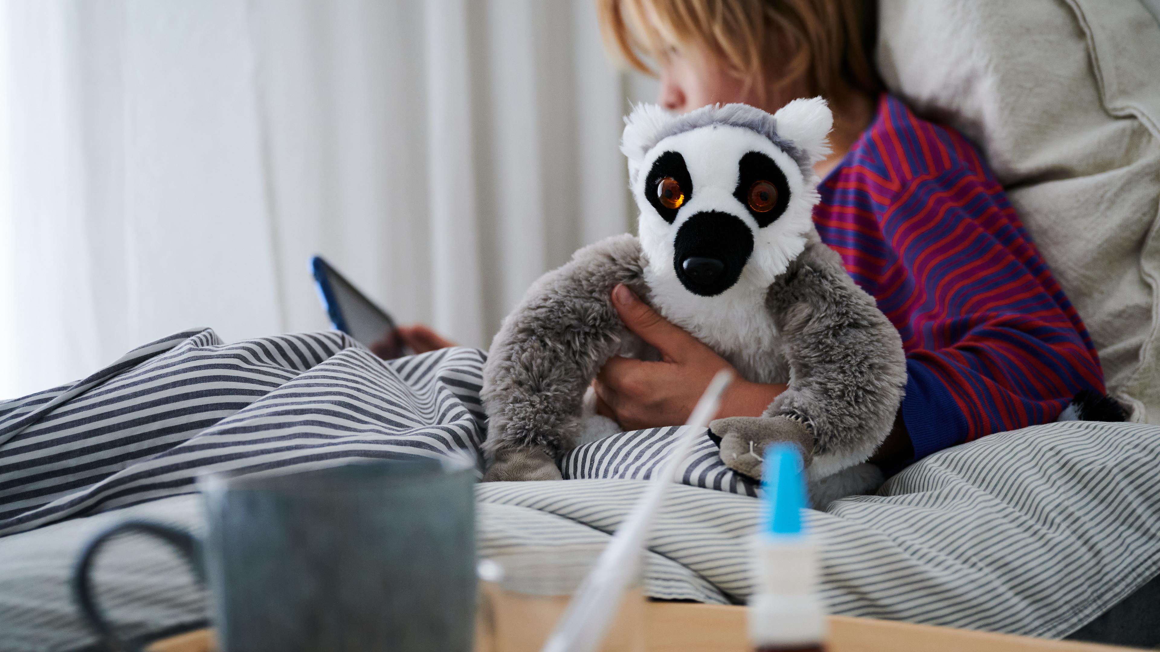 Ein Fieberthermometer, Nasenspray und eine Tasse Tee stehen vor einem Bett, in dem ein Kind mit Kuscheltier sitzt und auf ein Tablet schaut. Kleine Kinder sind oft krank. 