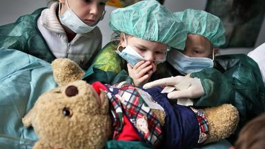 Drei Kindergartenkinder in klinischer Schutzkleidung und mit Maske sehen dabei zu, wie einem Teddybär im Vordergrund spielerisch der Blinddarm entfernt wird.