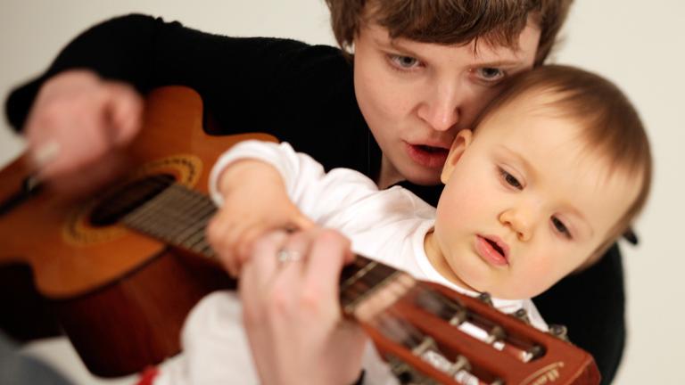 Eine Mutter hält ihr Kleinkind und eine Gitarre und spielt darauf.