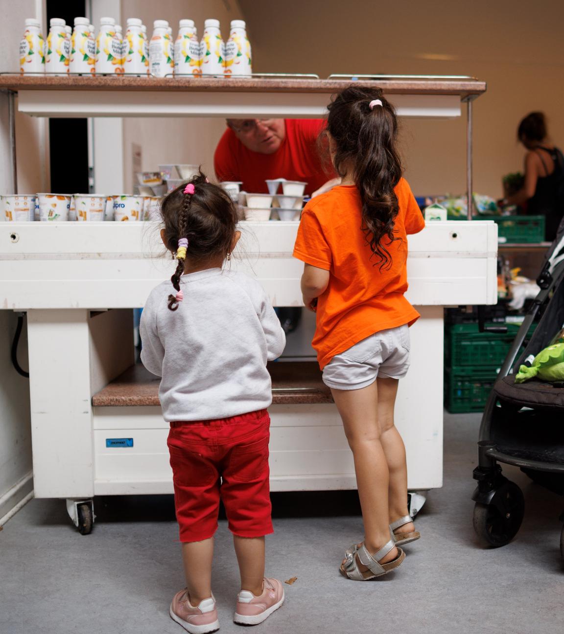 Ein Mitarbeiter verteilt Jogurt an zwei Kinder in der Ausgabestelle Paul-Schneider-Haus der Berliner Tafel. Kinder sind von hinten zu sehen.