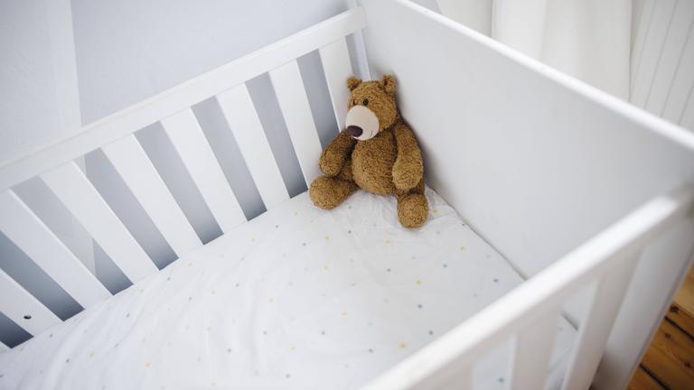 Ein Teddybaer sitzt in einem leeren Kinderbett. Symbolbild zum Thema unerfüllter Kinderwunsch.