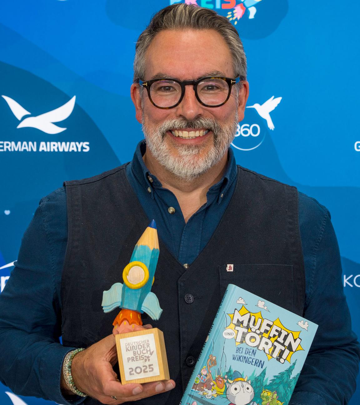 Der Autor Adam Stower hält sein Buch ""Muffin und Tört - Bei den Wikingern" und eine Preistrophäe in den Händen.
