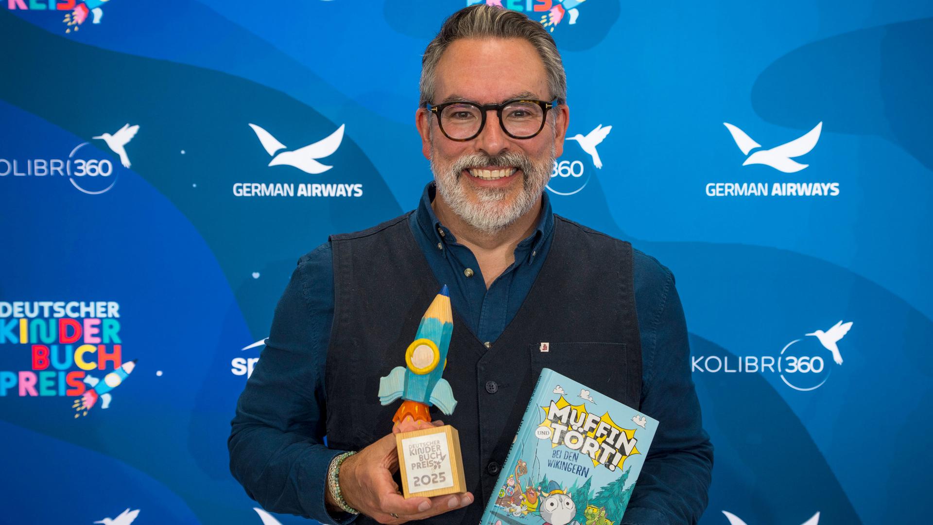 Der Autor Adam Stower hält sein Buch ""Muffin und Tört - Bei den Wikingern" und eine Preistrophäe in den Händen.