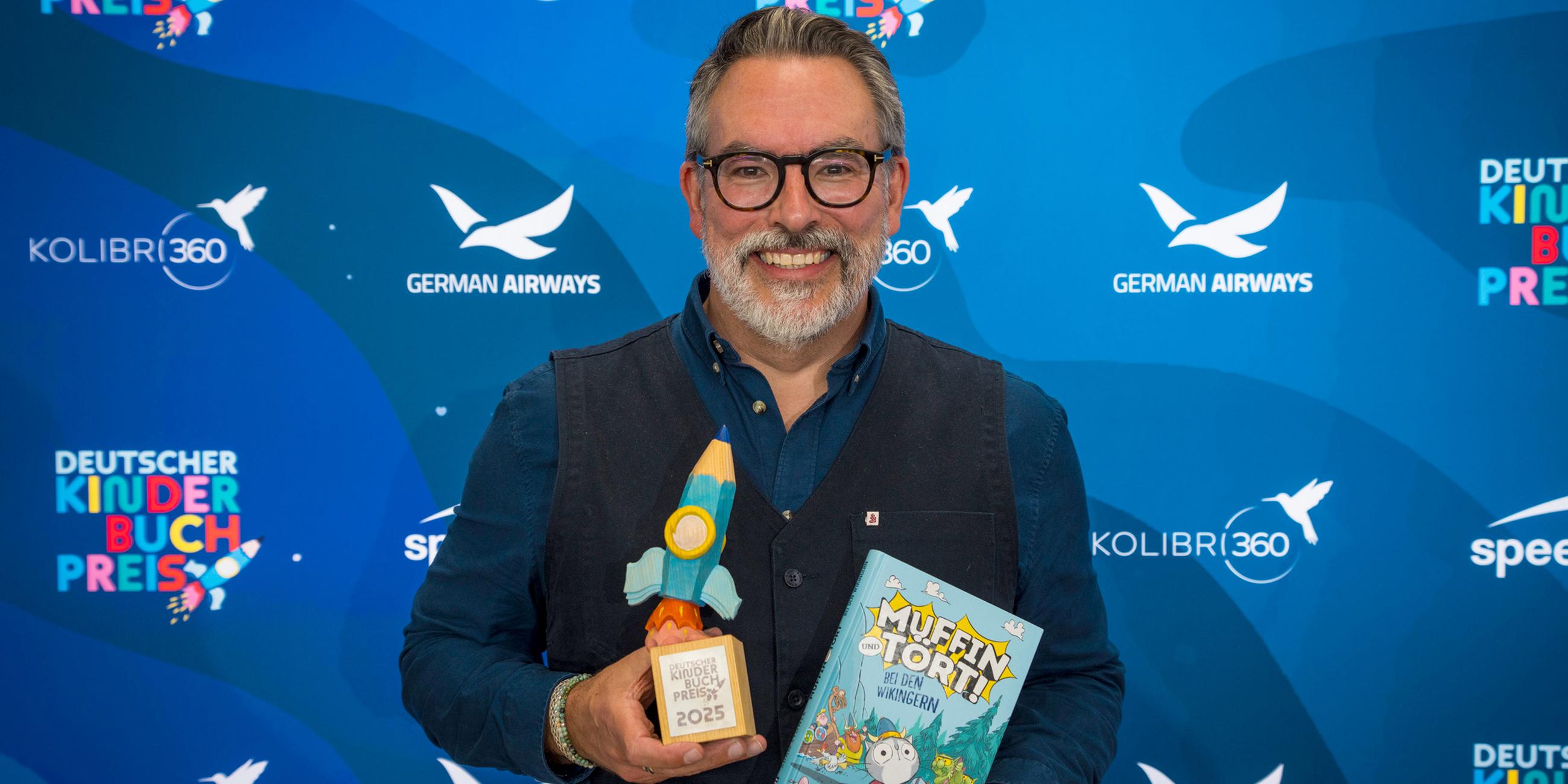 Der Autor Adam Stower hält sein Buch ""Muffin und Tört - Bei den Wikingern" und eine Preistrophäe in den Händen.
