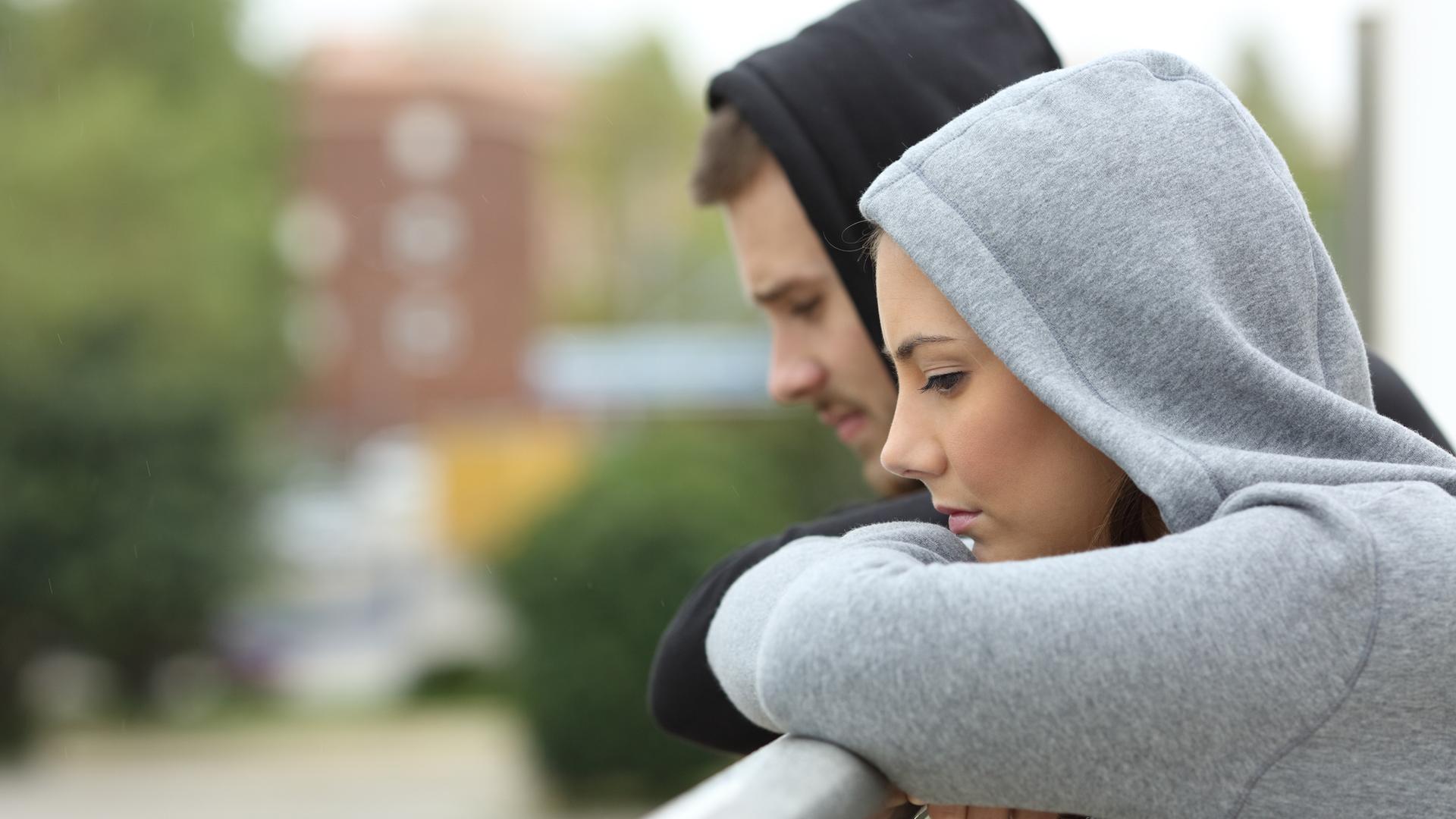 Eine junge Frau und ein junger Mann stehen auf einem Balkon und schauen traurig in die Ferne.