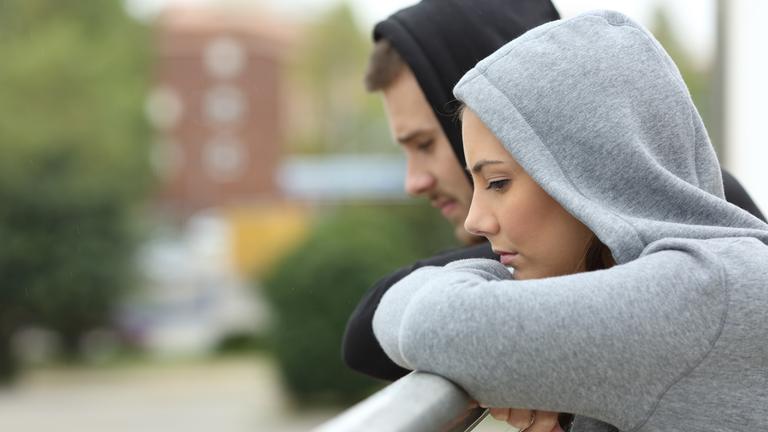 Eine junge Frau und ein junger Mann stehen auf einem Balkon und schauen traurig in die Ferne.