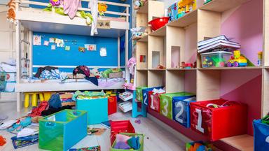 Völliges Durcheinander auf dem Boden von Spielzeug und Kleidung in einem Kinderzimmer mit Hochbett und Regalen.