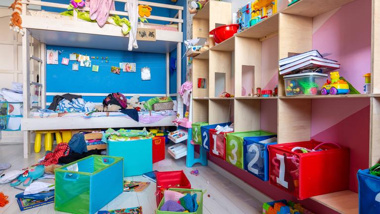 Völliges Durcheinander auf dem Boden von Spielzeug und Kleidung in einem Kinderzimmer mit Hochbett und Regalen.