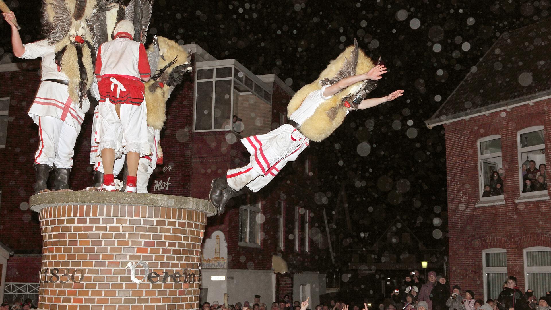 Niedersachsen, Borkum: Mit Masken verkleidete Männer des Vereins Borkumer Jungens stürzen sich am Abend in der Innenstadt der Nordseeinsel von einer Litfaßsäule in die Arme der Schaulustigen.