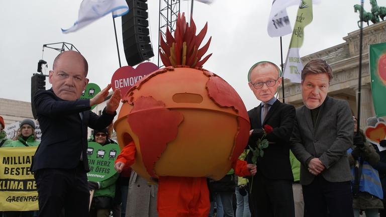 Karrikaturen von Scholz, Merz und Habeck beim Klimademo vor dem Brandenburger Tor