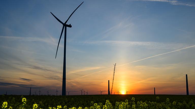 29.04.2025, Brandenburg, Lebus: Der Sonnenaufgang leuchtet über der Landschaft mit Windenergieanlagen und einem Kran auf einer Baustelle für ein neues Windrad.