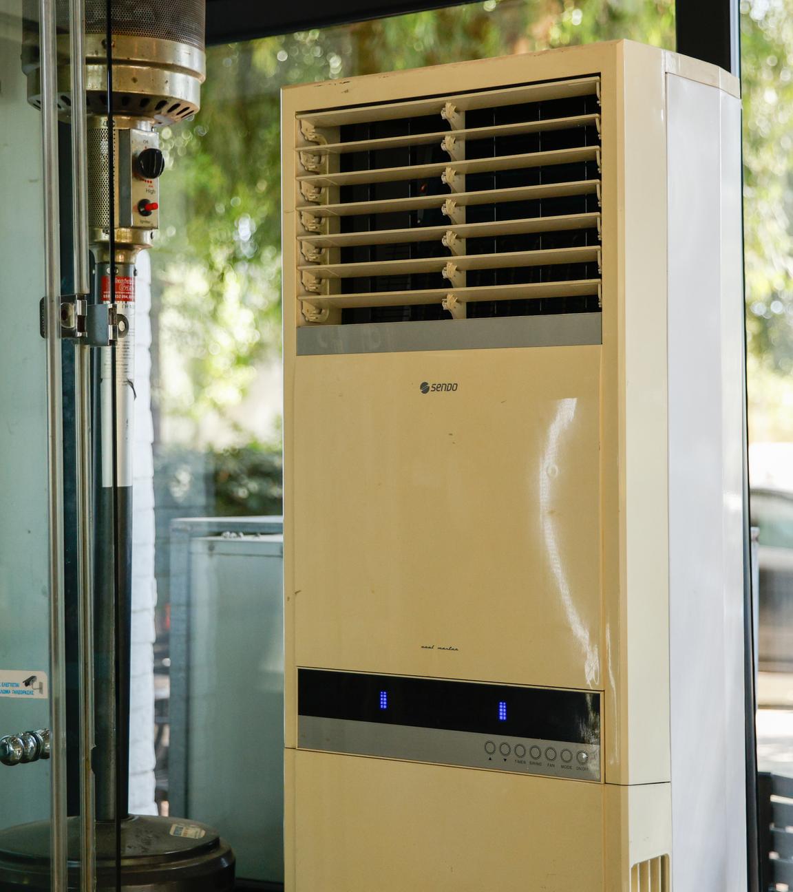 Griechenland, Athen: Eine mobile Klimaanlage steht in einem verglasten Innenraum eines Cafés.