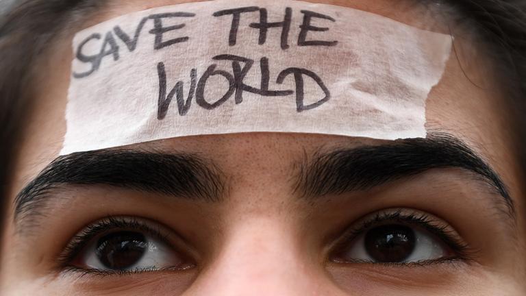 Ein Mädchen mit braunen Haaren hat ein weißes Klebeband auf der Stirn. Darauf steht: Save the world.