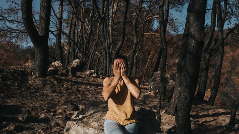 Symboilbild: Eine verzweifelte Frau, die ihr Gesicht verdeckt und auf einem Felsen in einem verbrannten Wald sitzt,