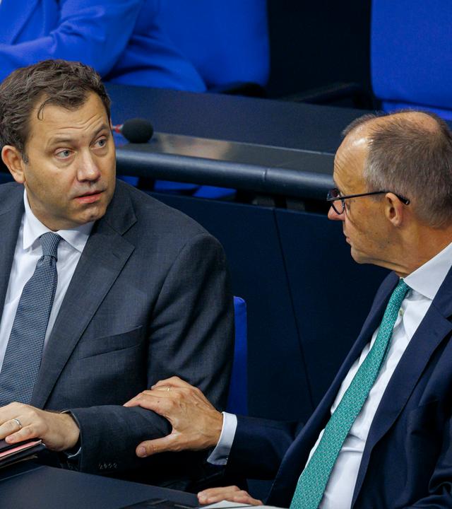 SPD-Chef Lars Klingbeil und Bundeskanzler Friedrich Merz (CDU) sitzen im Bundestag nebeneinander und sprechen miteinander.