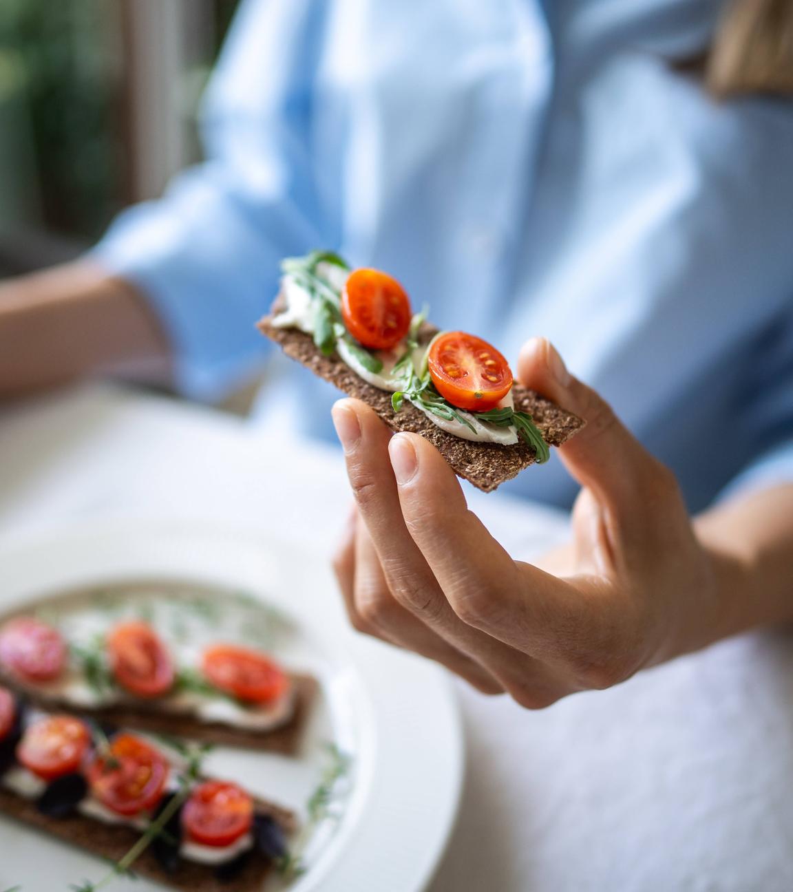 Eine Frau hält ein Knäckebrot in der Hand, das mit Käse, Tomaten und Basilikum belegt ist.