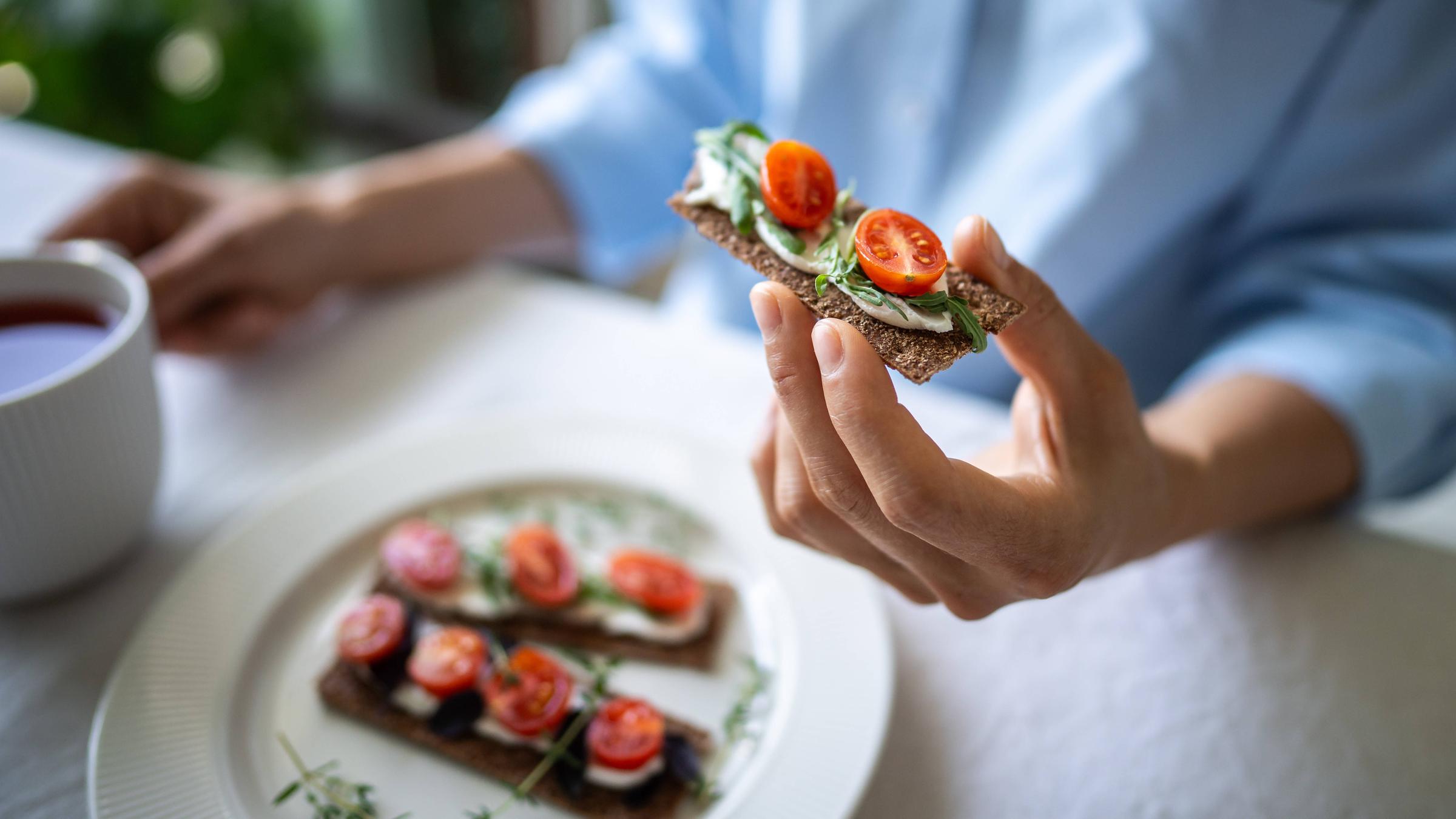 Eine Frau hält ein Knäckebrot in der Hand, das mit Käse, Tomaten und Basilikum belegt ist.