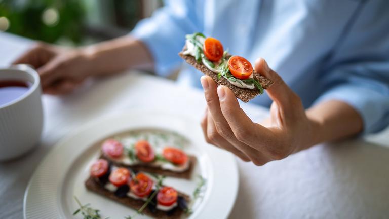 Eine Frau hält ein Knäckebrot in der Hand, das mit Käse, Tomaten und Basilikum belegt ist.