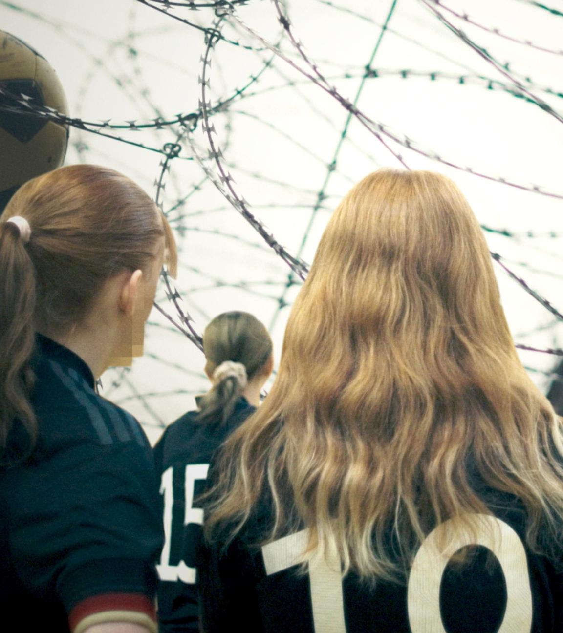 Junge Gefängnisinsassinnen in Fußballtrikots; im Hintergrund ein Ball im Stacheldraht