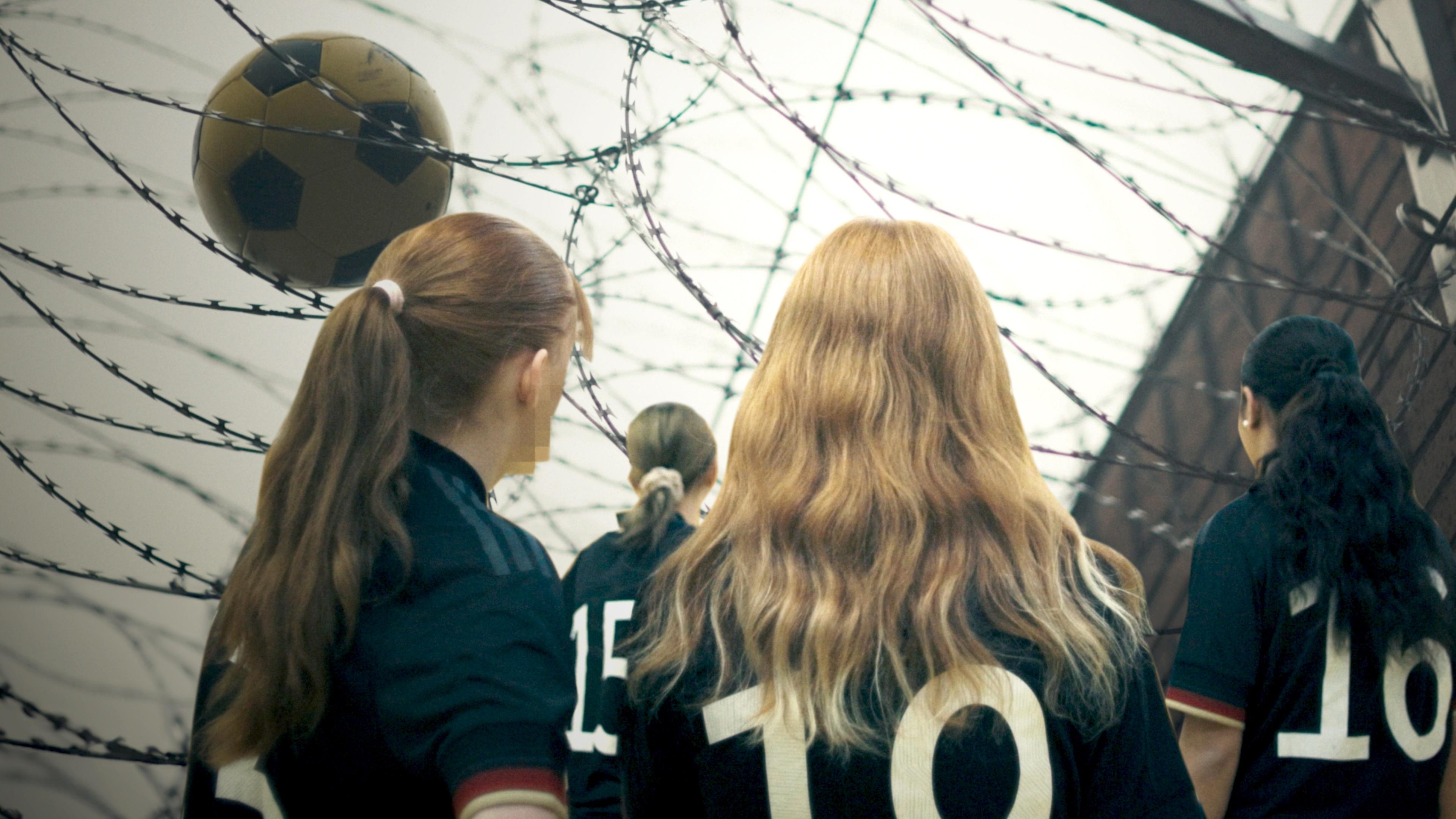 Junge Gefängnisinsassinnen in Fußballtrikots; im Hintergrund ein Ball im Stacheldraht