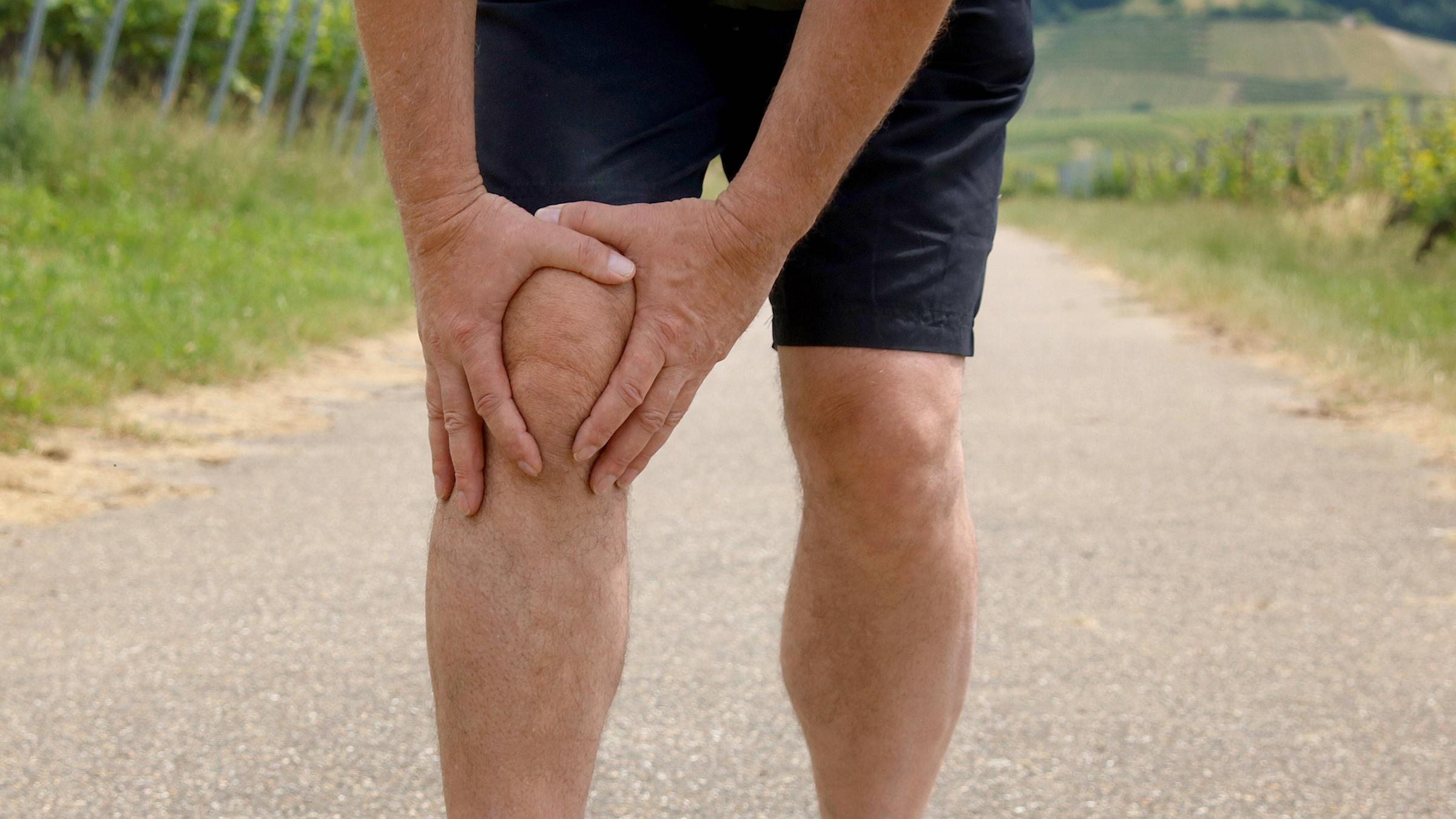 Ein Jogger in weißen Schuhe und schwarzer, kurzer Hoser hält sich sein schmerzendes Knie.