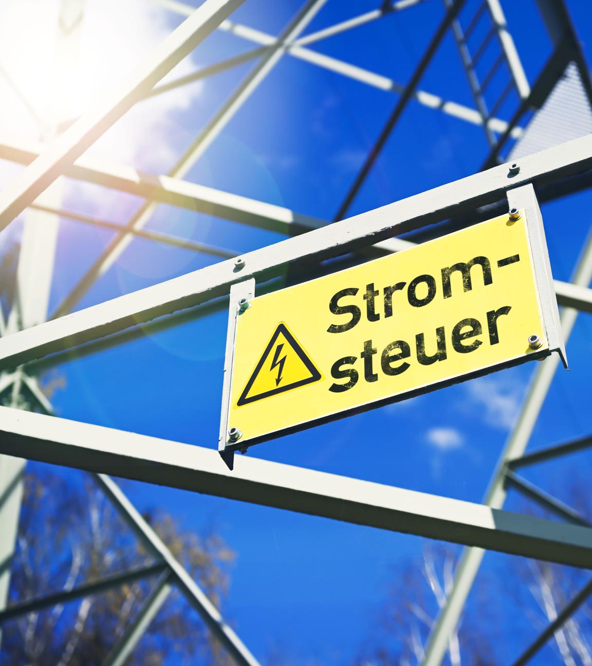 Ein gelbes Schild mit der Aufschrift "Stromsteuer" neben einem gelben Blitz der auf Hochspannung hinweist hängt an einer Stromleitung.