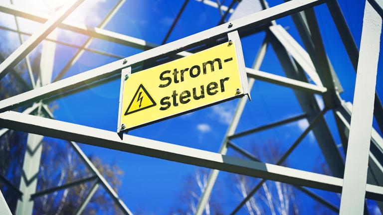 Ein gelbes Schild mit der Aufschrift "Stromsteuer" neben einem gelben Blitz der auf Hochspannung hinweist hängt an einer Stromleitung.