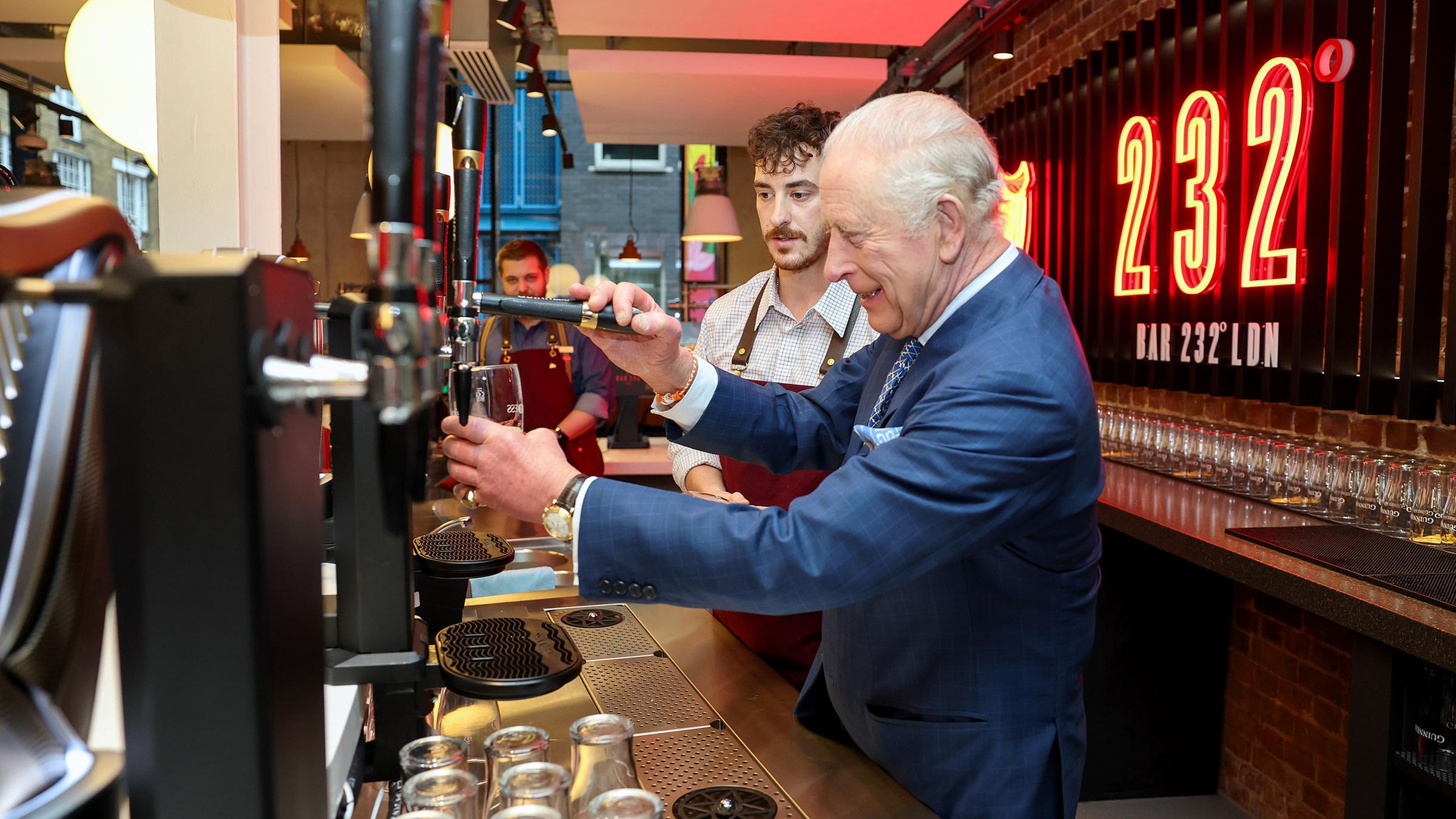 König Charles steht hinter einem Tresen und zapft ein Bier in ein Bierglas.