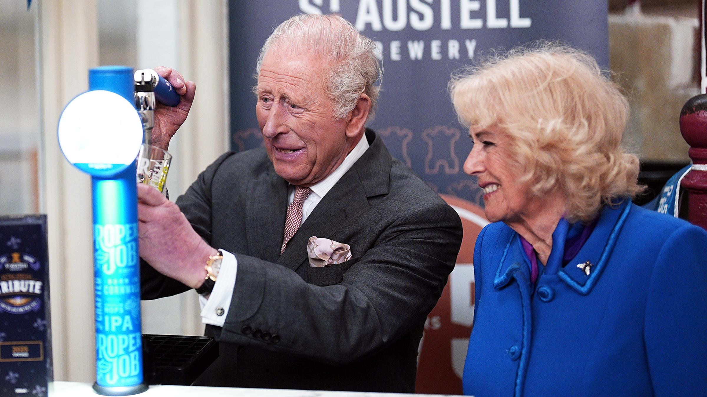 König Charles (links) zapft ein Bier, seine Frau Camilla steht neben ihm und freut sich darüber. 