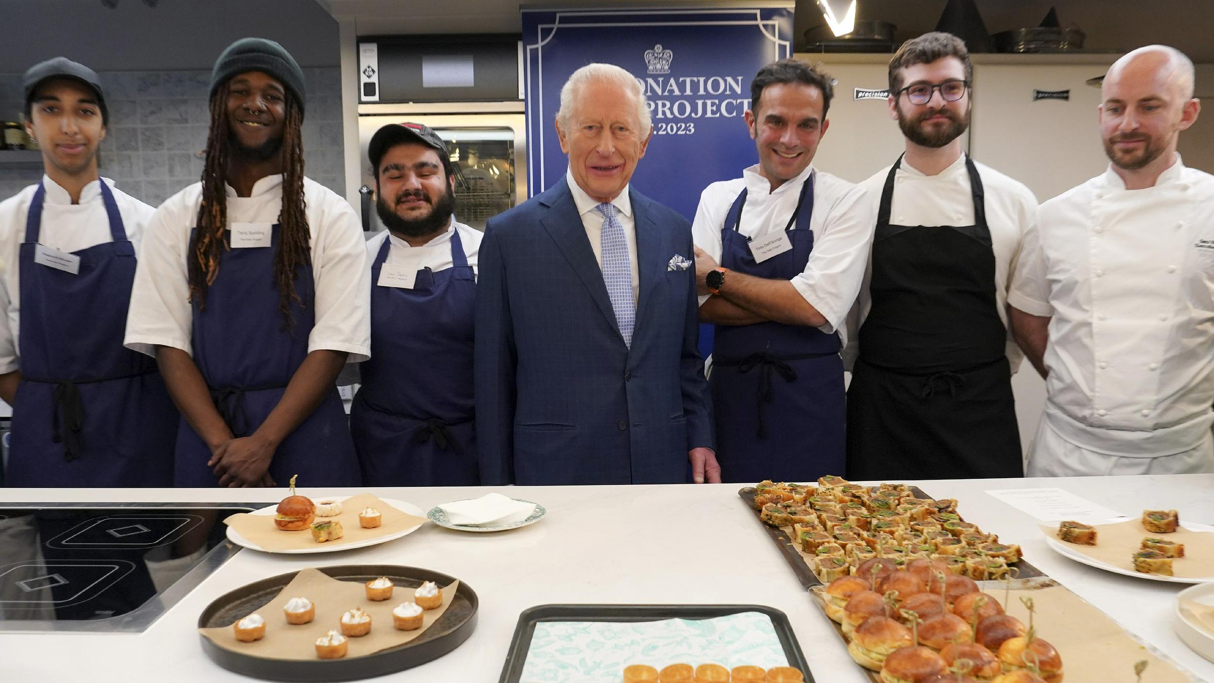 Der britische König Charles steht in einem dunkelblauen Anzug zusammen mit sechs Köchen vor einer Auswahl an kleinen Speisen.