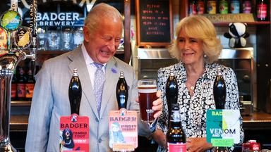 König Charles (rechts) zapft ein Bier, neben ihm steht seine Frau Königin Camilla.