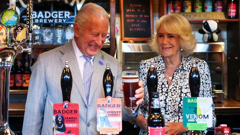 König Charles (rechts) zapft ein Bier, neben ihm steht seine Frau Königin Camilla.