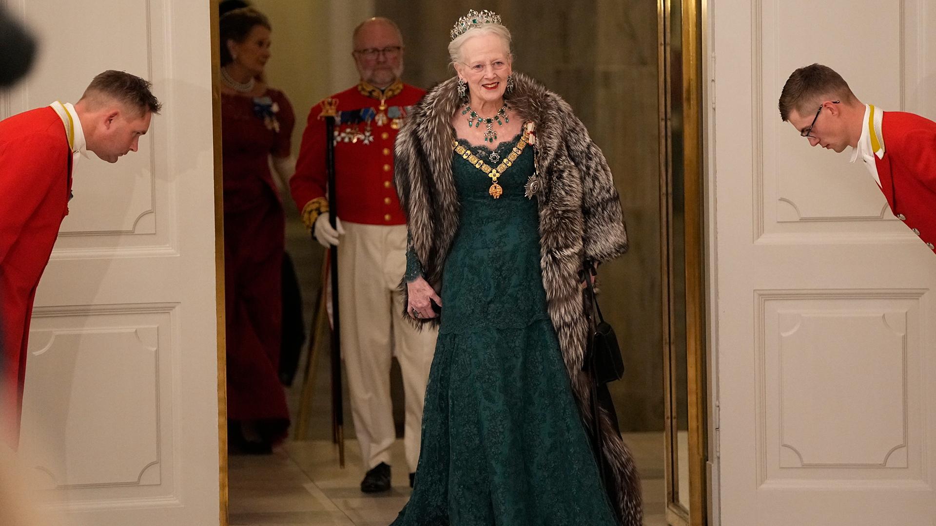 Königin Margrethe II. tritt in einem grünen Kleid durch eine Tür bei einem Staatsbankett im Schloss Christiansborg. Rechts und Links befinden sich zwei Angestellte, die sich vor der Königin verbeugen. Im Hintergrund befinden sich weitere Gäste.