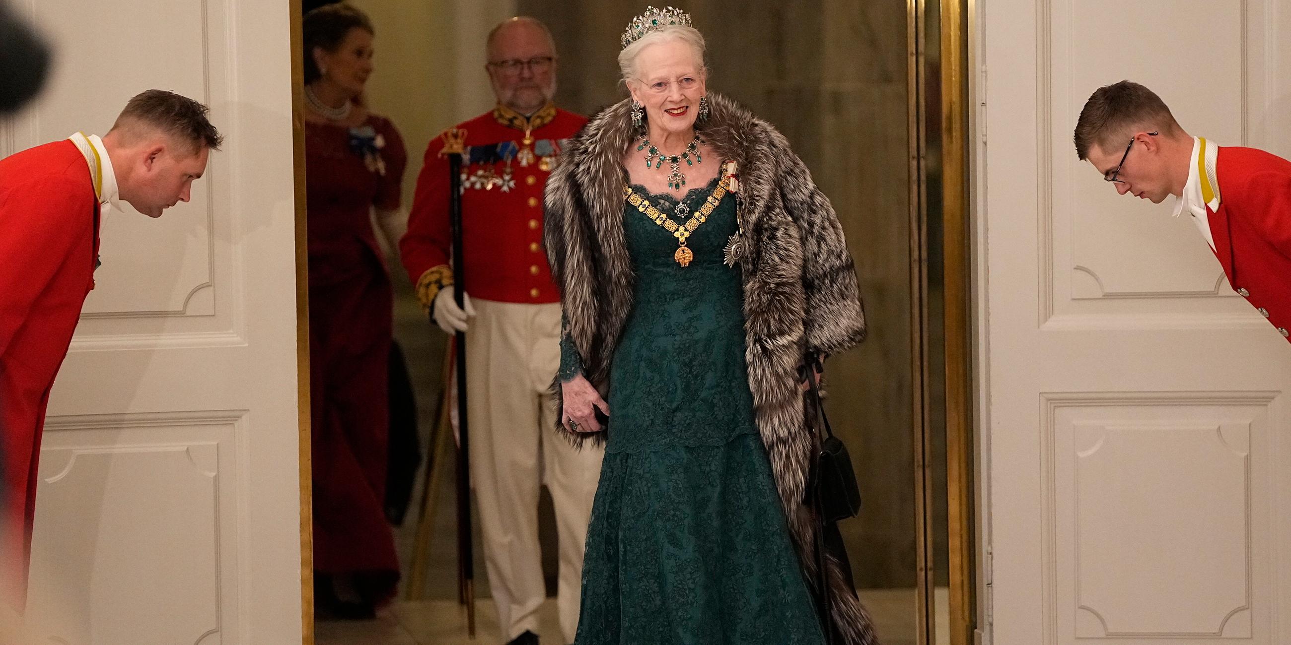 Königin Margrethe II. tritt in einem grünen Kleid durch eine Tür bei einem Staatsbankett im Schloss Christiansborg. Rechts und Links befinden sich zwei Angestellte, die sich vor der Königin verbeugen. Im Hintergrund befinden sich weitere Gäste.