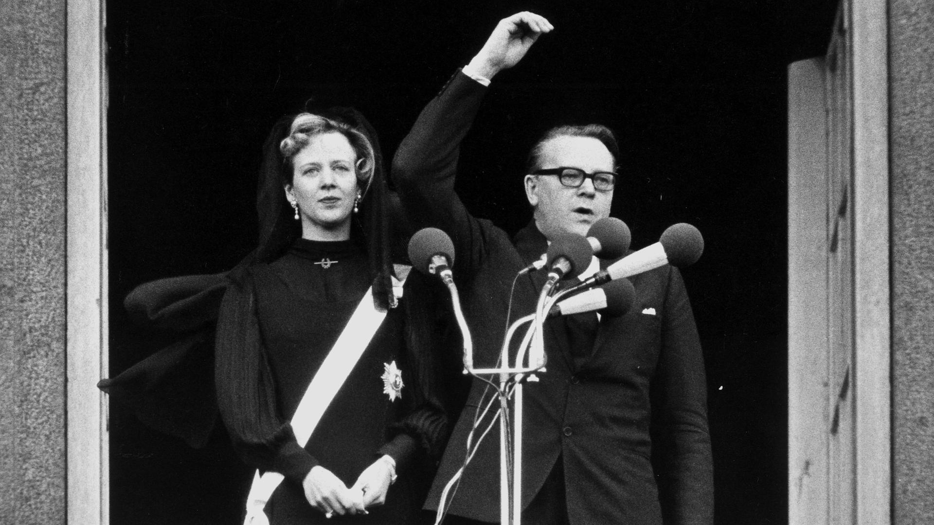 Ein schwarz-weiß Bild aus dem Jahre 1972. Zu sehen ist Margrethe II. von Dänemark, die neben dem Premierminister Jens Otto Krag auf einem Balkon steht und zur Königin ernannt wird.