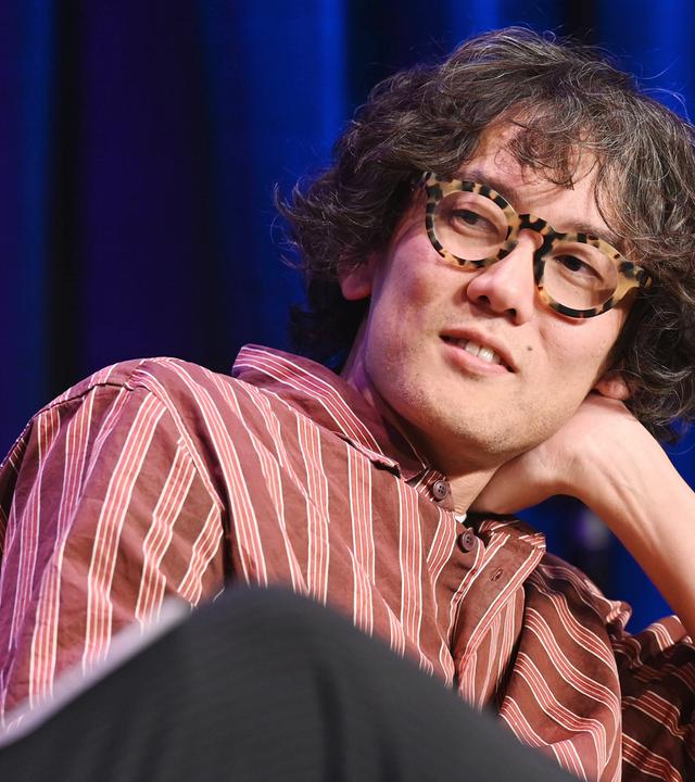 Kohei Saito - ein Japaner mit kurzen leicht graumelierten lockigen Haaren und großer Kunststoffbrille stützt sich auf eine Hand im Nacken, während er einer Person, die neben ihr sitzt, zuhört.