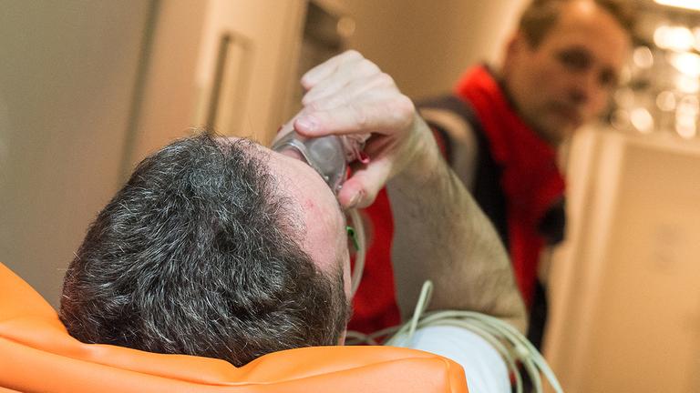 Ein Patient liegt auf einer Trage und trägt eine Sauerstoffmaske. Er wird von einem Sanitäter geschoben.