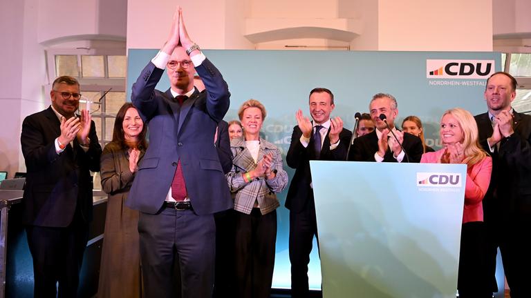 Ministerpräsident Hendrik Wüst ( CDU ) hebt die Arme nach seinem Statement. Im Hintergund ist das Logo und weitere Vertreter der CDU.