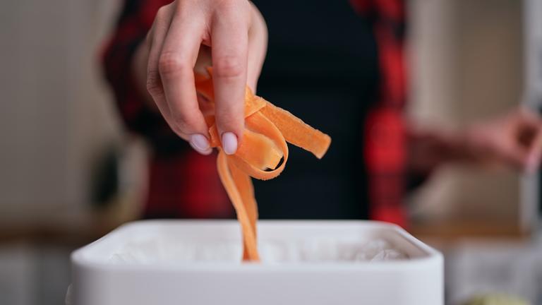 Eine Hand wirft Möhrenschale in einen weißen Eimer.