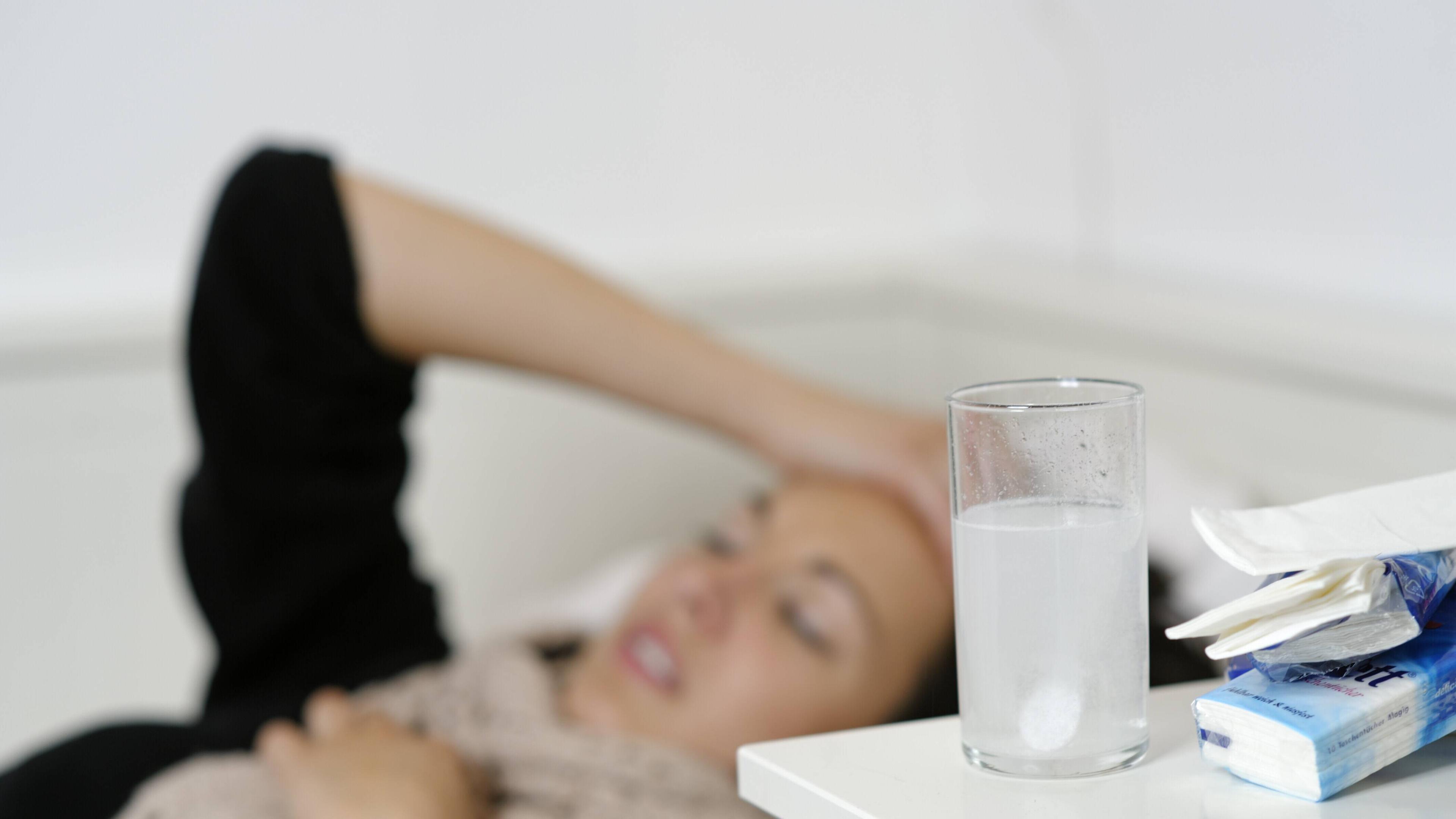 Junge Frau liegt im Bett und fasst sich an den Kopf. Vor ihr steht ein Glas mit einer Tablette. 