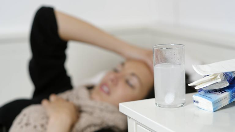 Junge Frau liegt im Bett und fasst sich an den Kopf. Vor ihr steht ein Glas mit einer Tablette. 