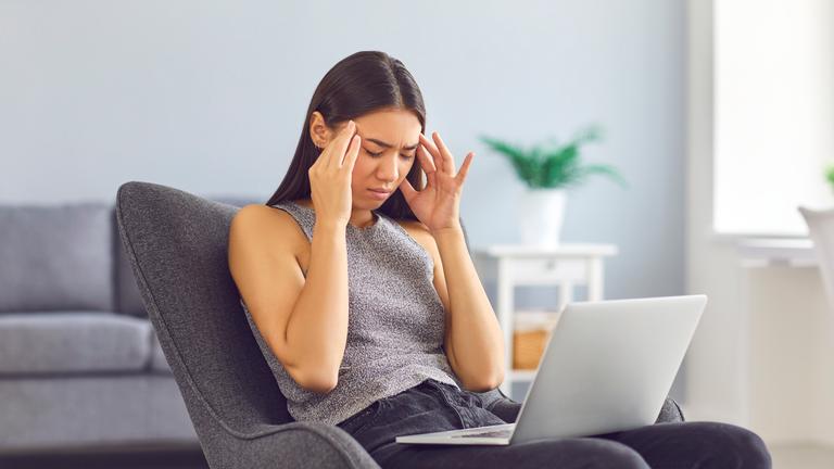 Eine Frau sitzt mit ihrem Laptop im Schoß auf einem Stuhl und massiert ihre Schläfen.